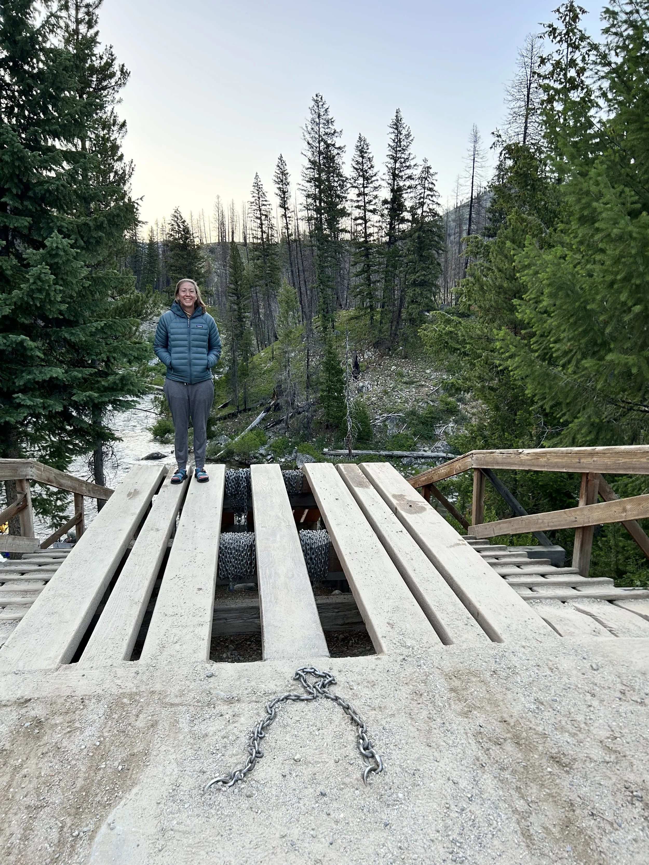 A person standing on a wooden bridge in a forested area near a river, with tall pine trees and mountains in the background, during daylight.
