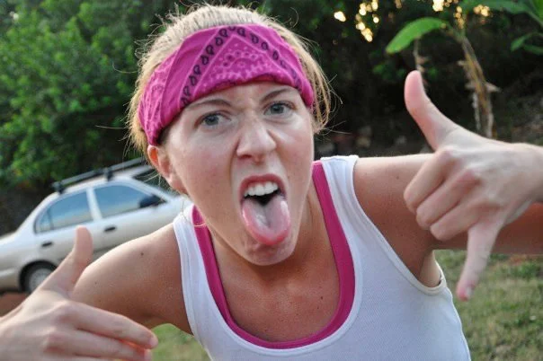 A woman wearing a pink bandana and white tank top making a silly face with her tongue out, giving a thumbs-up with both hands outdoors.