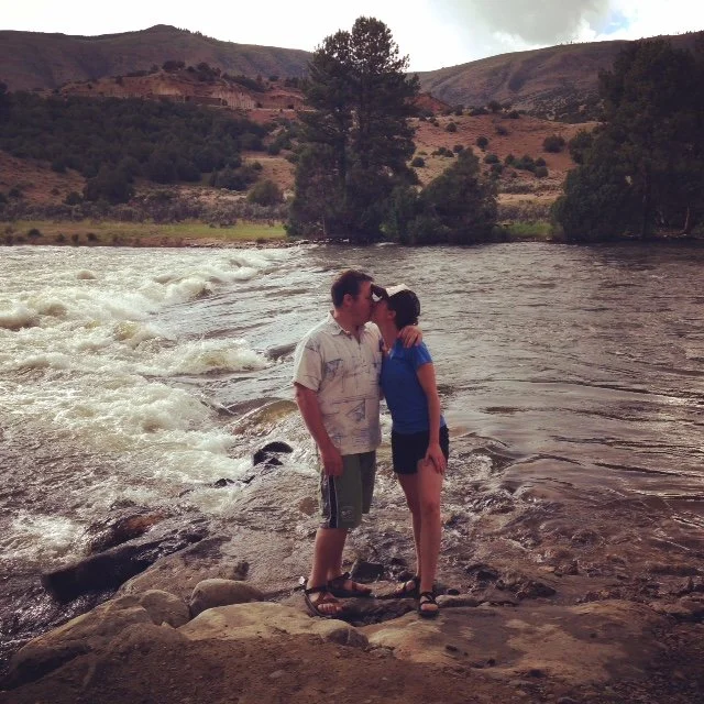 A couple stands on rocks by a river, kissing, with hills and trees in the background.
