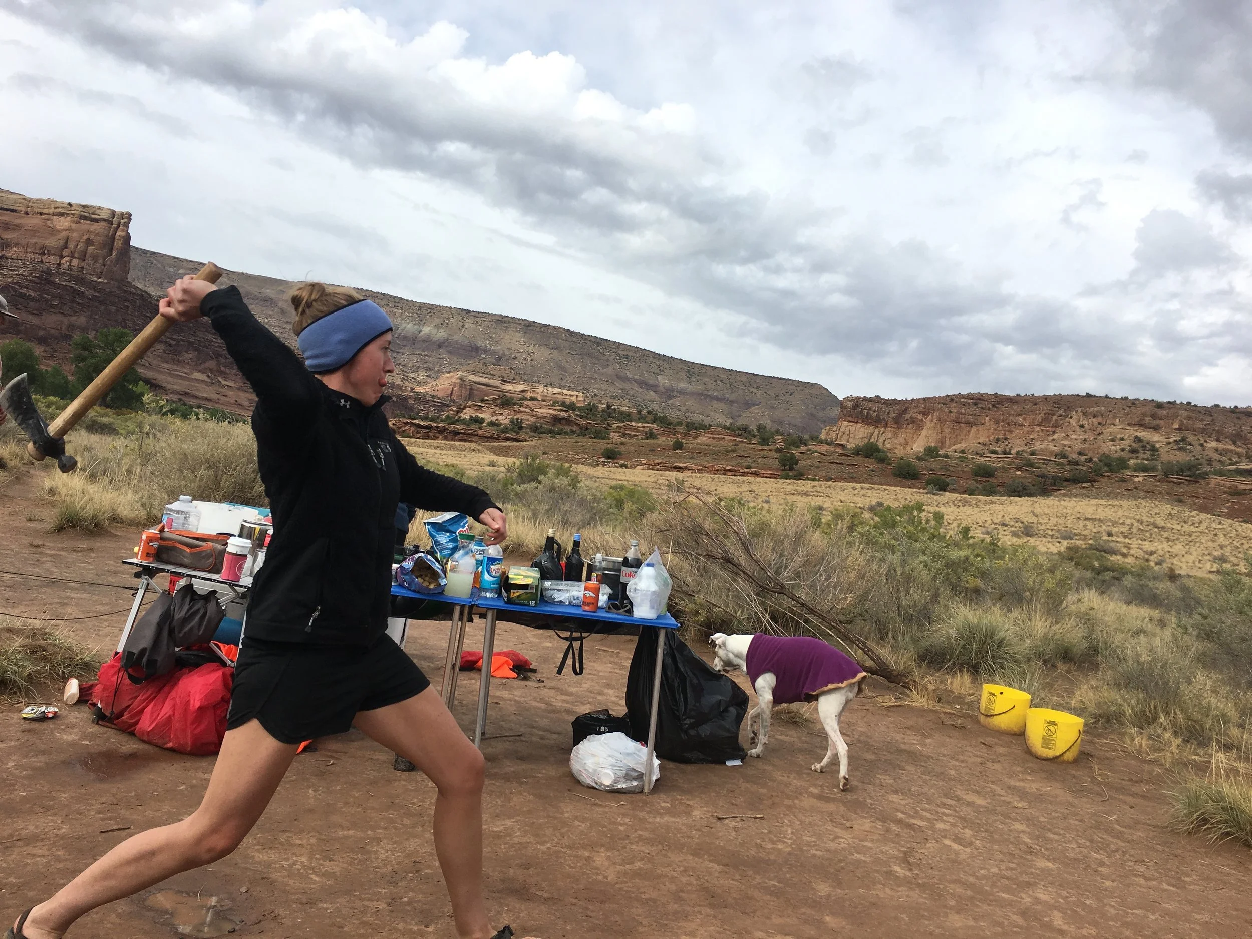 A woman in black athletic clothing and a headband swinging an axe in a desert landscape with a dog in a purple sweater and a table with supplies.