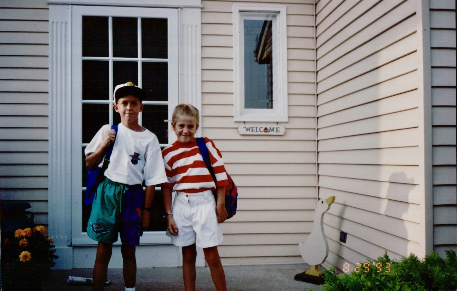 Two children standing outside a house with beige vinyl siding, a window, and a small decorative duck figure. The boy is wearing a white t-shirt, green shorts, a baseball cap, and a backpack, while the girl is wearing a red and white striped t-shirt, 