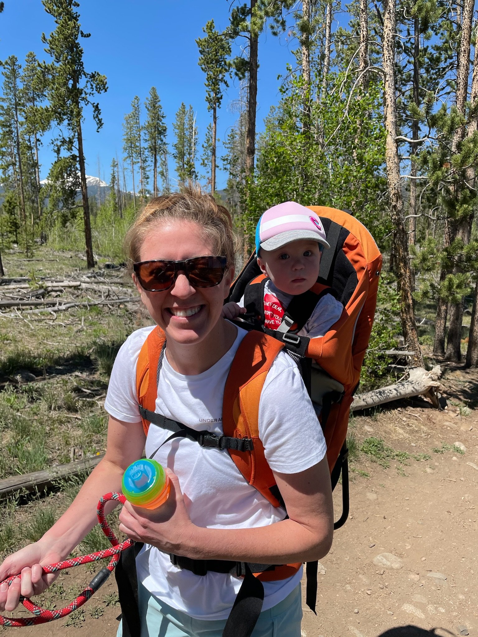 Smiling woman with sunglasses hiking in a forest, carrying a young child in an orange backpack carrier under a clear blue sky.