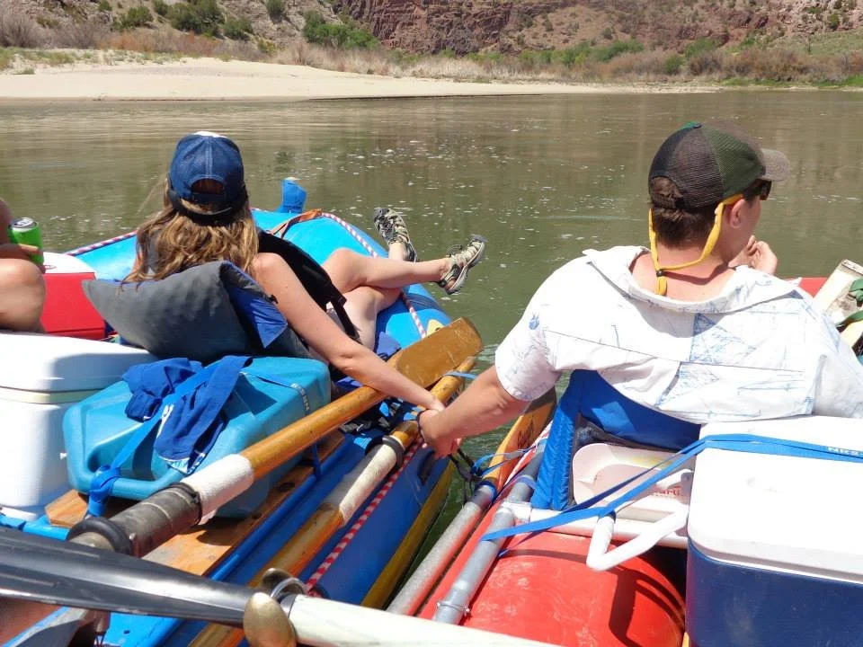 Two people on a raft in a river, with a sandy shoreline and rocky cliffs in the background. They are wearing casual clothing and hats, and appear to be relaxing or reading.