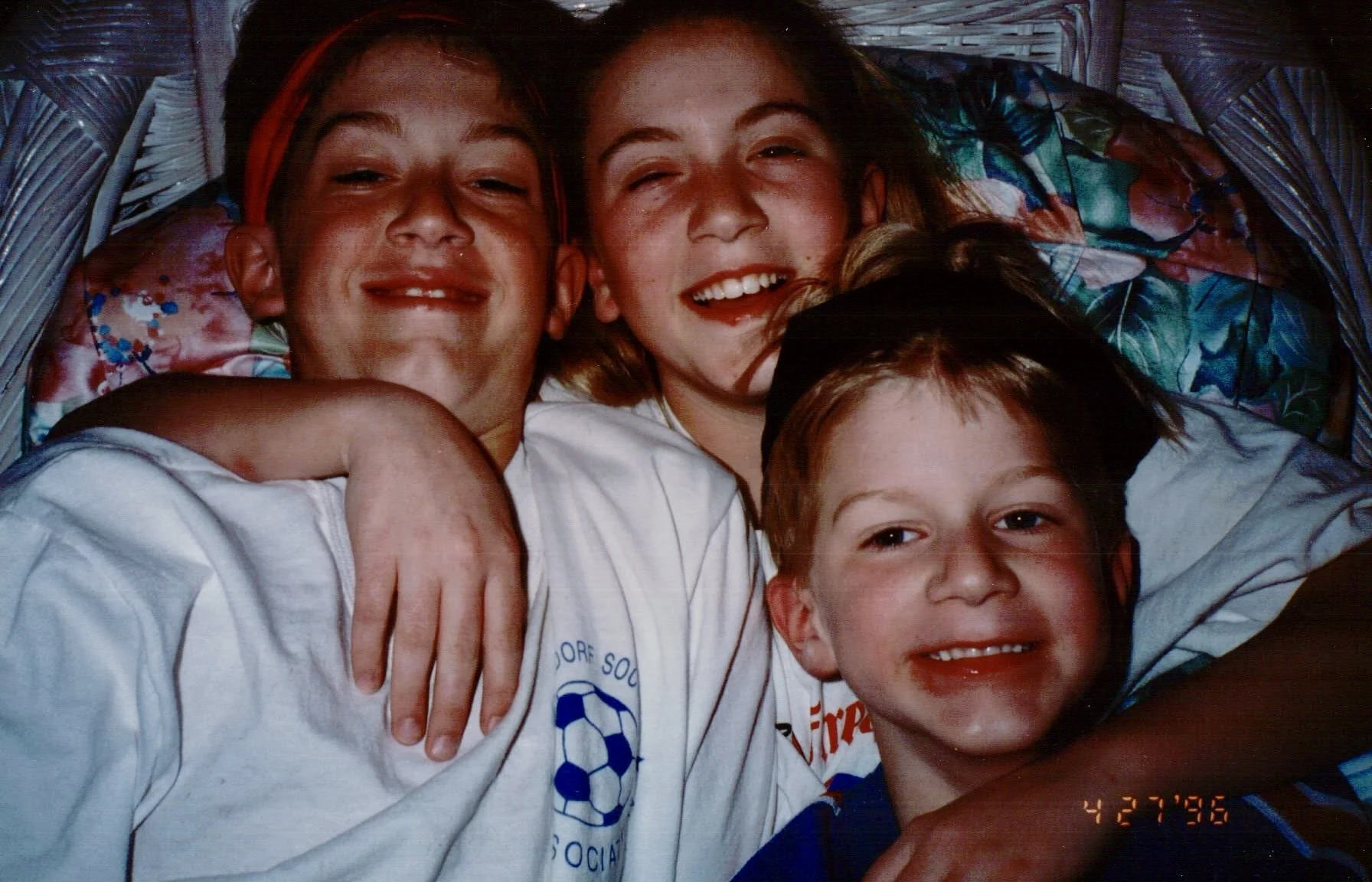 Four children happily laying together in a basket, wearing sports uniforms and one wearing a graduation cap, smiling and hugging each other.