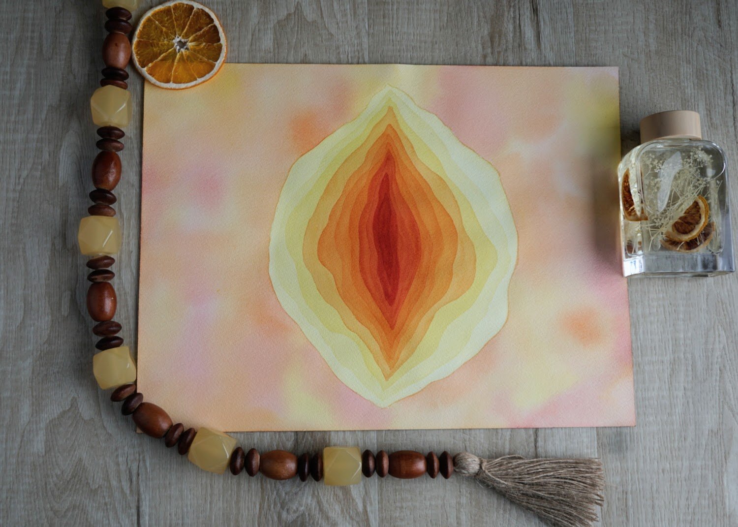 Flat watercolor painting with orange, yellow, and cream-colored symmetrical abstract pattern. Object on black background, dried orange slices in a glass bottle, and a beaded necklace with cream, brown, and wooden beads with a tassel are surrounding it.