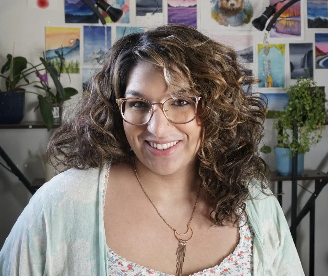 A woman with curly brown hair and glasses smiling at the camera. In the background, there are wall art and potted plants.