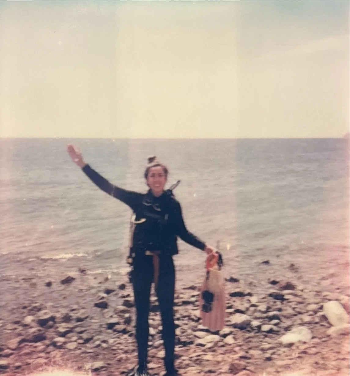 A woman scuba diver standing on a rocky beach near the water, smiling and waving with her right hand, holding diving fins in her left hand, wearing a dark wetsuit.