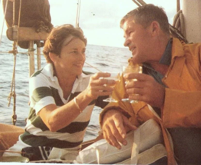 Vintage photo of two people on a sail boat sharing a toast with drinks, smiling at each other, with the ocean in the background.