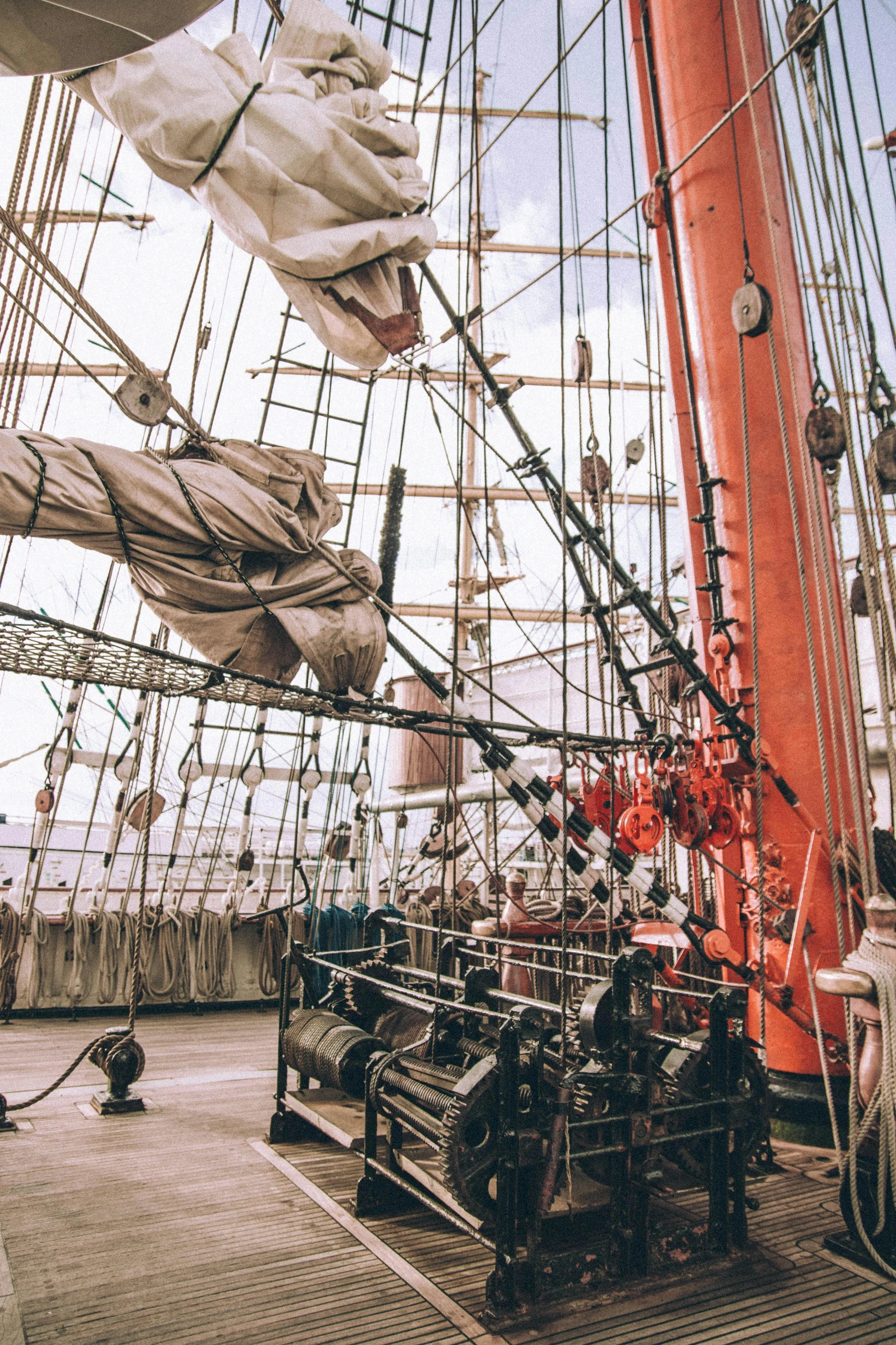Close-up of the deck and rigging of a historic sailing ship, showing folded sails, ropes, pulleys, and parts of the wooden deck with a reddish-orange mast and rigging.