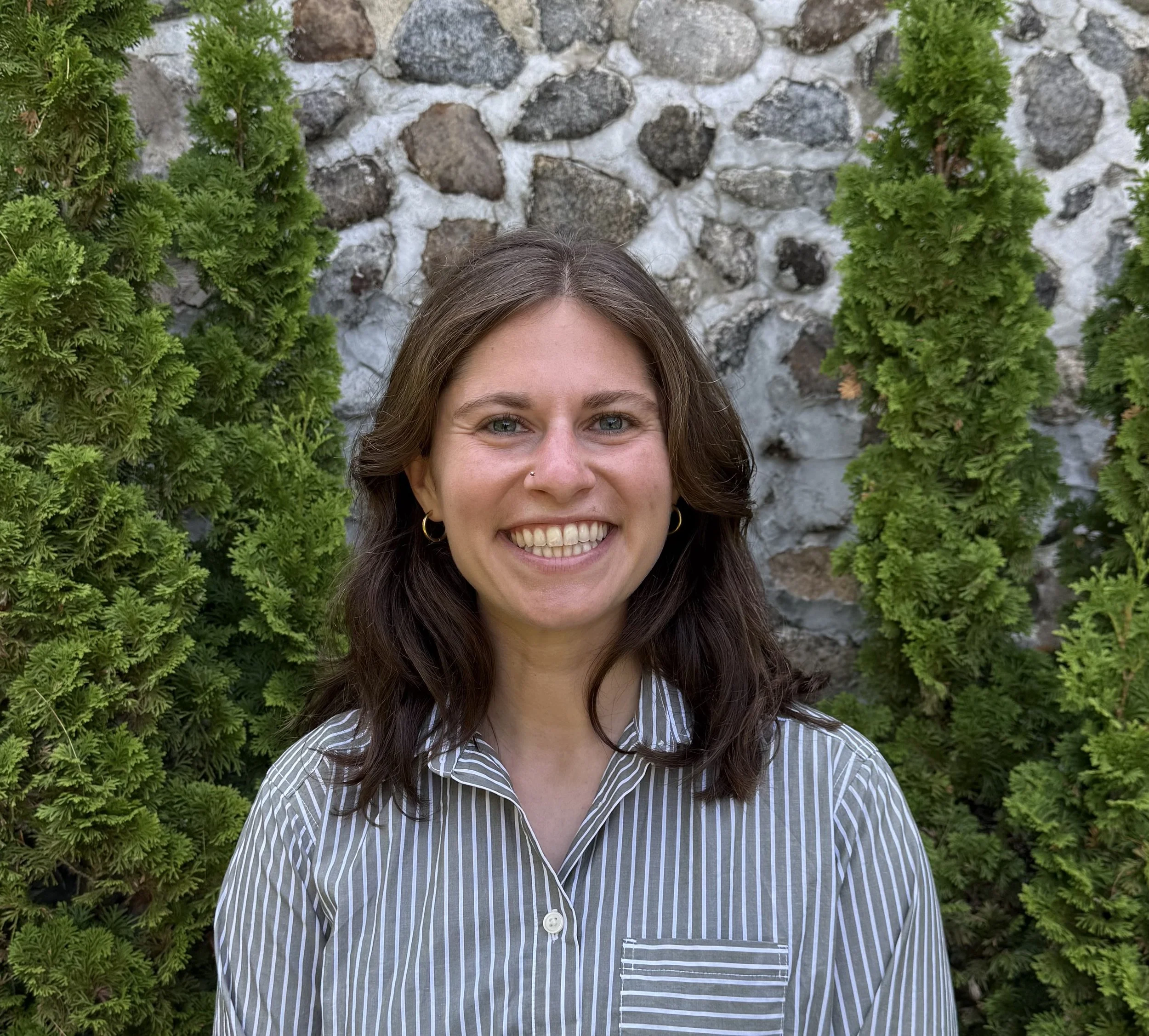 Smiling woman with dark wavy hair, wearing a striped button-up shirt and hoop earrings, standing outdoors in front of a stone wall with green bushes.