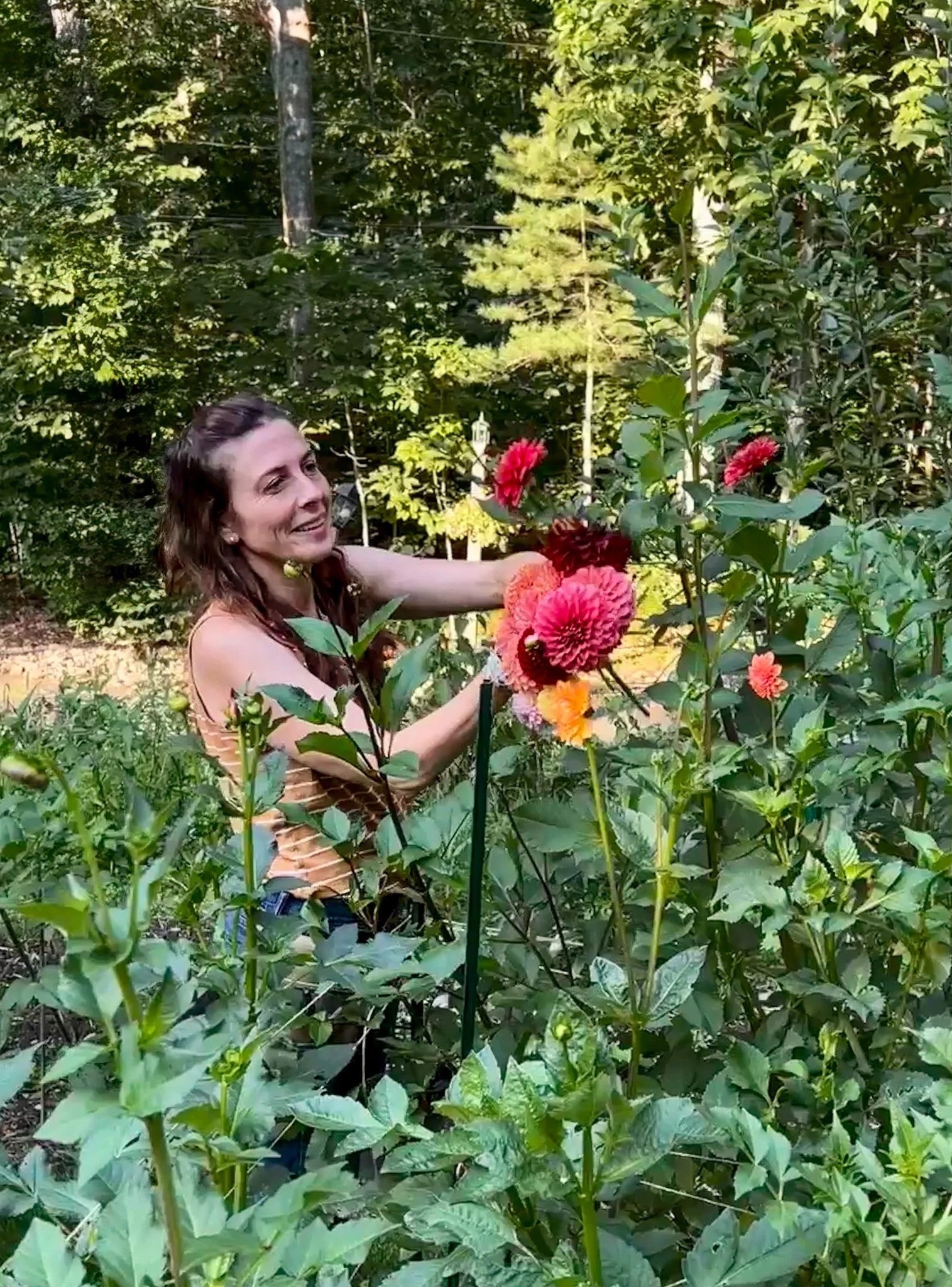 A woman in an outdoor garden with green foliage and trees, smiling and picking pink and orange flowers.