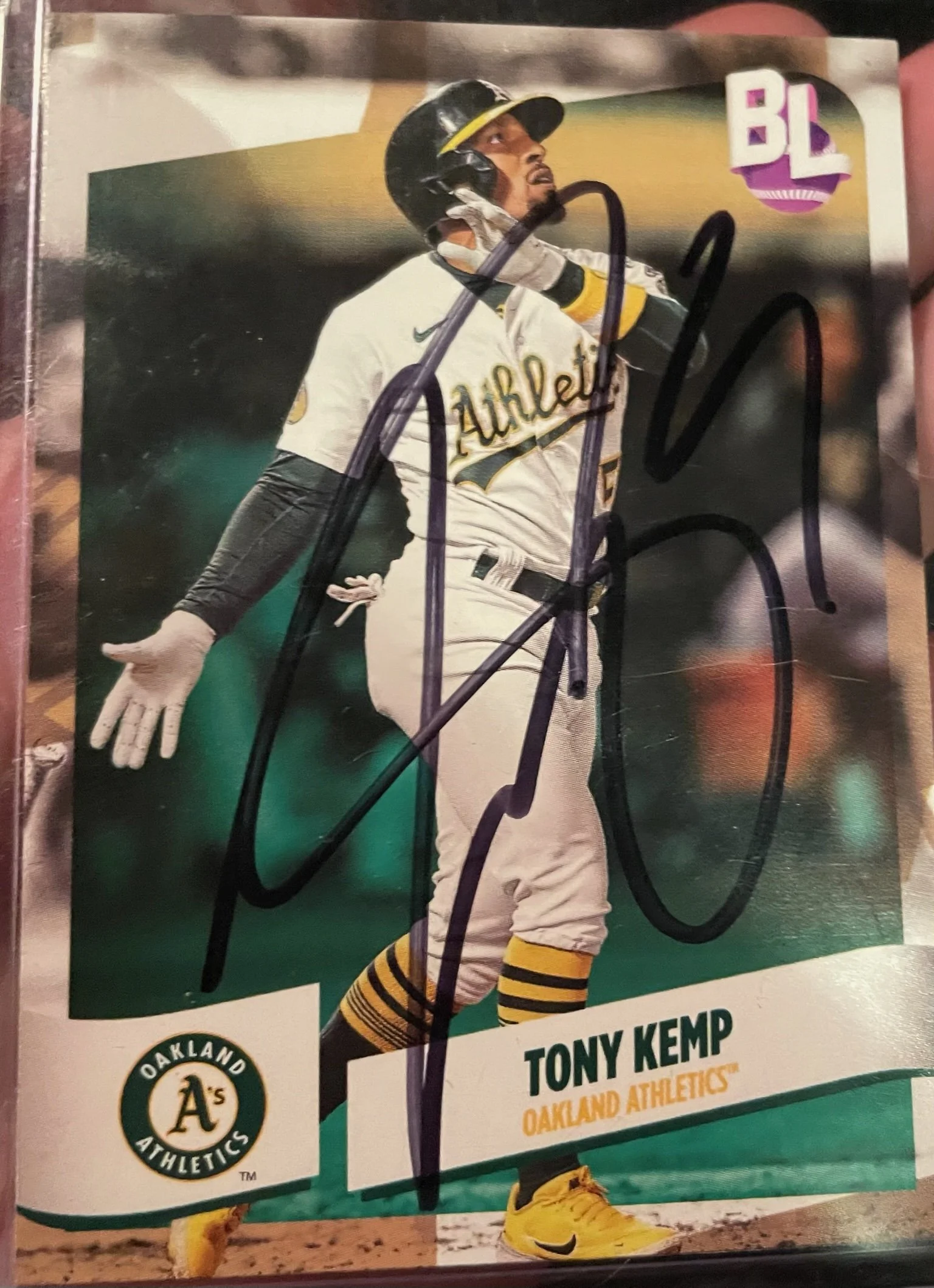 Autographed baseball card of Tony Kemp from Oakland Athletics, featuring him in a batting pose wearing the team uniform and black helmet, with black and yellow socks, with a large black signature across the front.