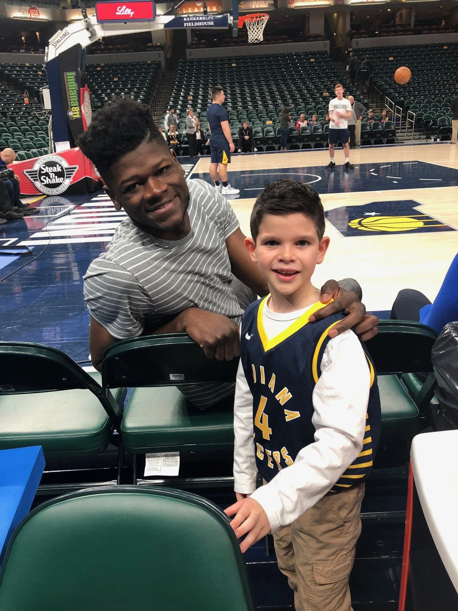 Two young boys at a basketball game, one sitting on a green seat and the other leaning on the seat back, in an arena with a court in the background. The court has players practicing and seating visible behind.