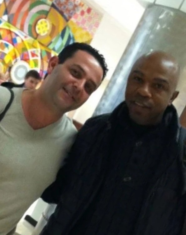 Two men smiling, one with light skin and short dark hair wearing a light-colored shirt, and the other with dark skin and a shaved head wearing a dark jacket, standing in front of a colorful abstract art piece.