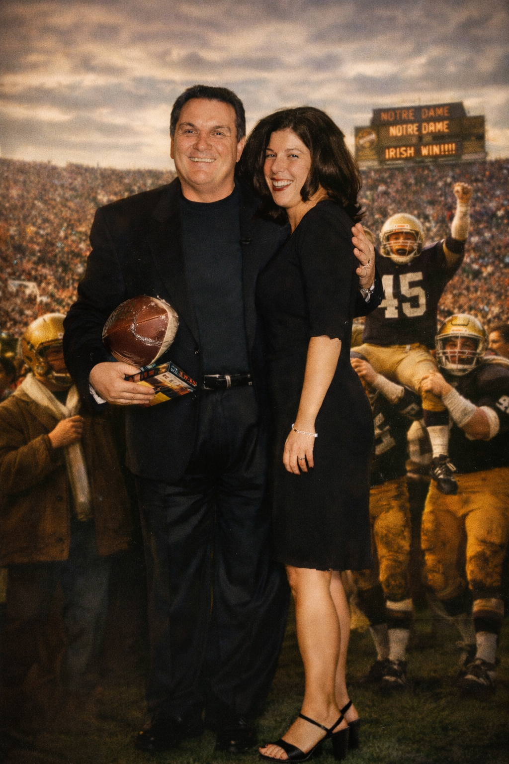 A man and a woman standing together at a football game, smiling at the camera. The man holds a football, and there is a celebratory sign in the background that says 'Notre Dame Irish Win!!!!'