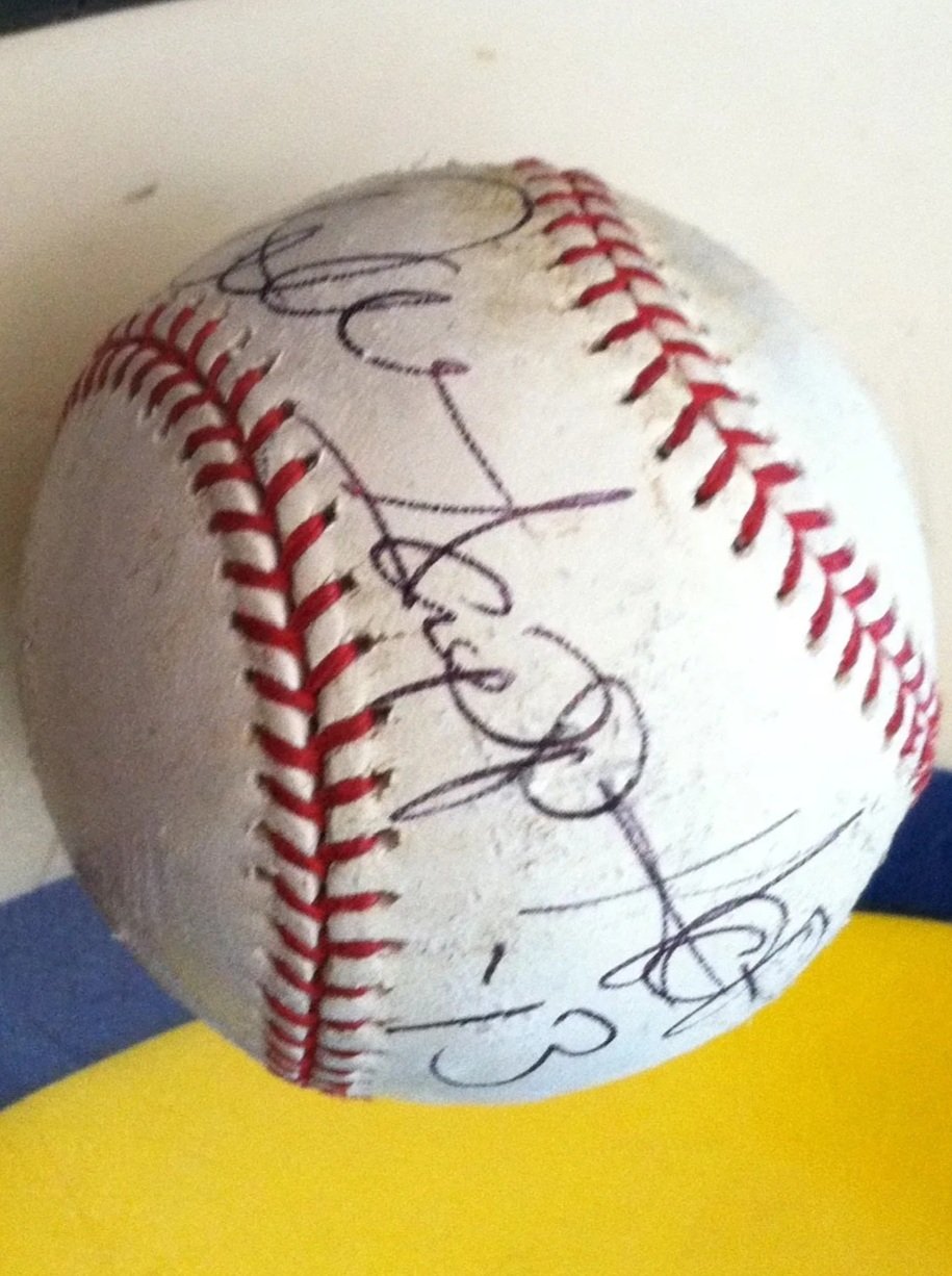 A white baseball with red stitching and black autograph signatures on its surface, resting on a multicolored yellow, black, and blue surface.
