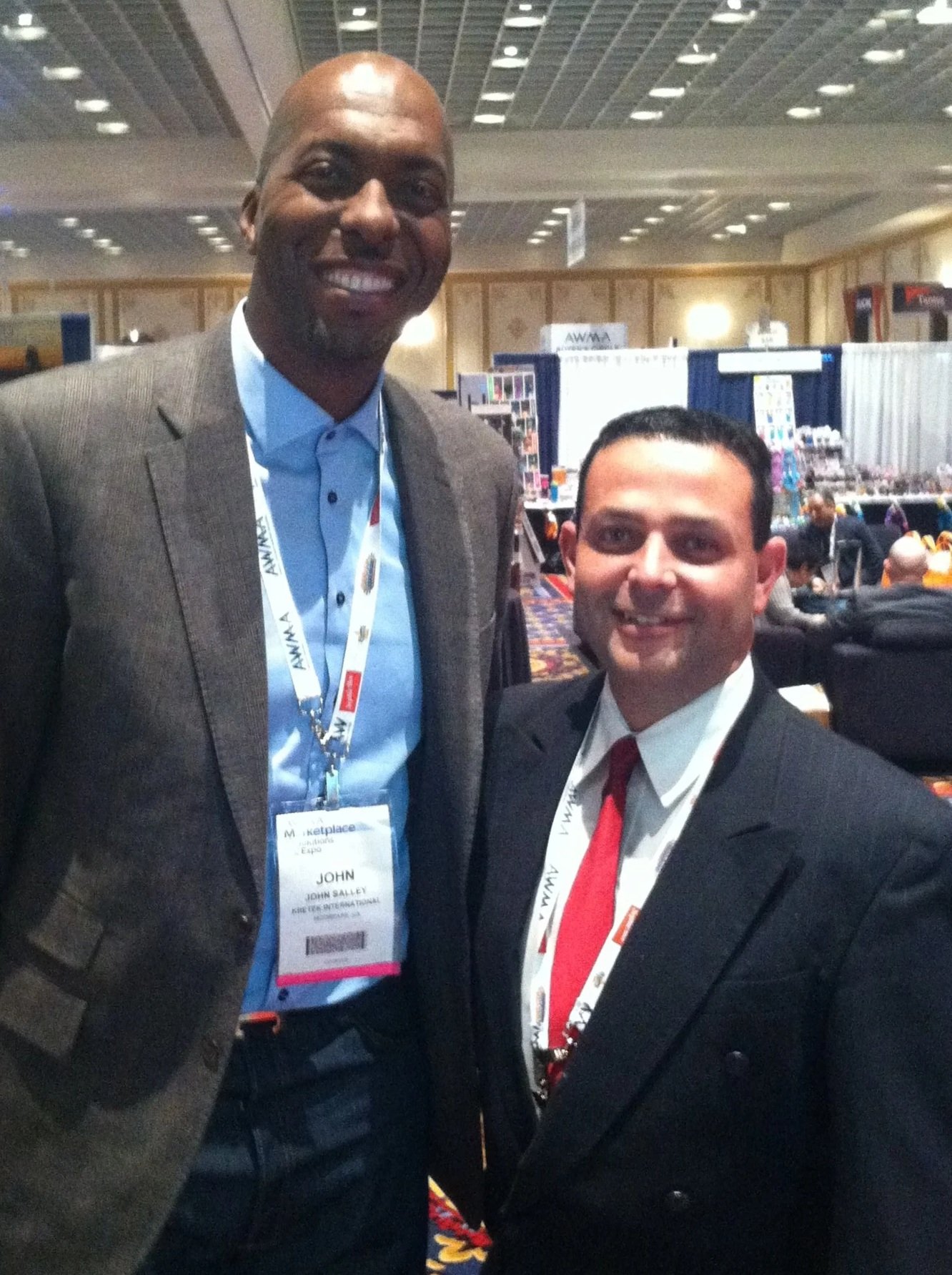 Two men dressed in suits at a conference, smiling, with conference badges around their necks, standing in a large ballroom.
