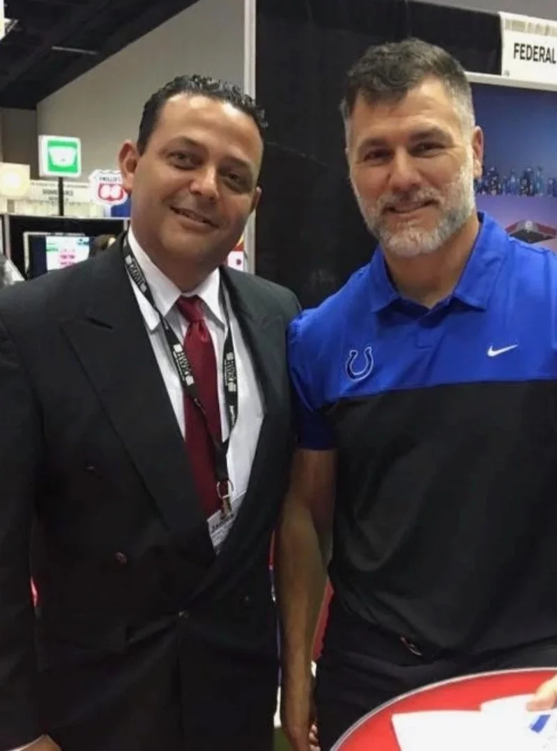 Two men standing side by side at an indoor event, smiling at the camera. One is wearing a suit with a tie, and the other is dressed in a blue and black sports shirt with a Colts logo.