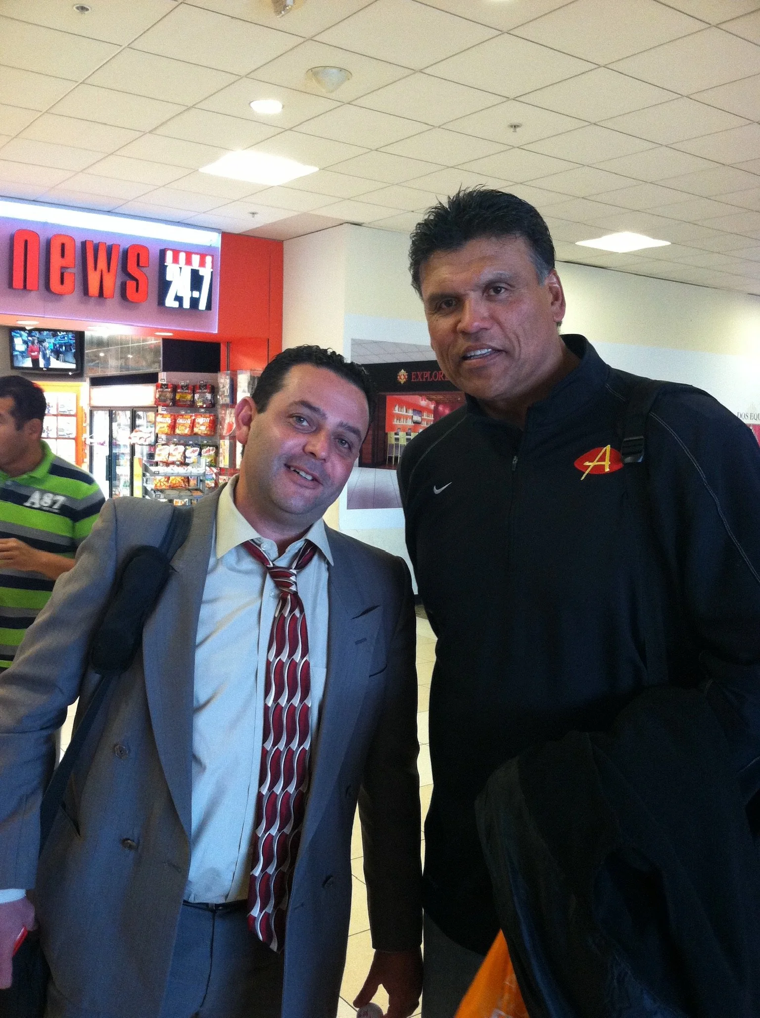 Two men posing for a photo in an airport, one in a suit and the other in athletic casual wear, with a newsstand in the background.