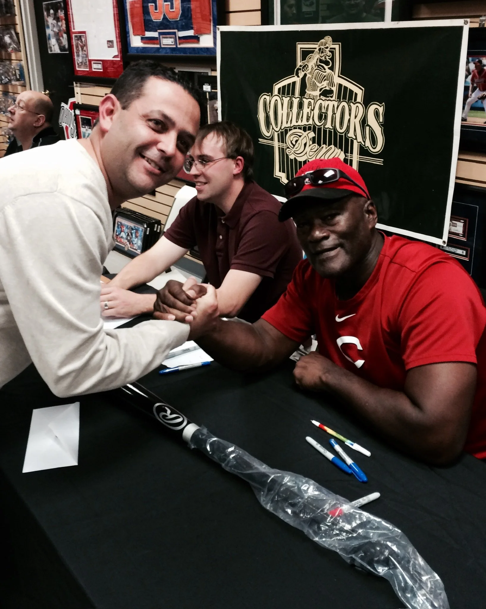 Two men, one in a beige shirt and the other in a red shirt with a Superman logo, arm wrestling and shaking hands across a table at a sports memorabilia signing event, with a third man in the background and a banner in the background that reads 'Colle