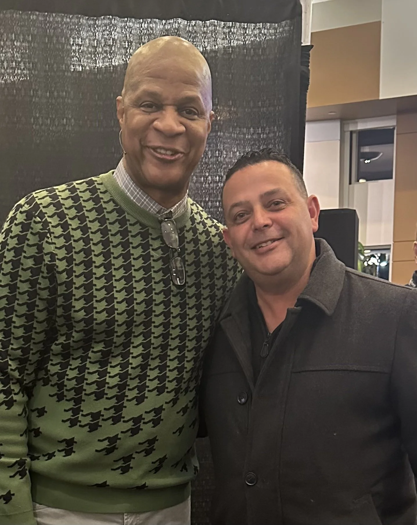 Two men standing close together indoors, smiling at the camera. One man is wearing a patterned sweater and glasses hanging from his collar, and the other is dressed in a black coat.