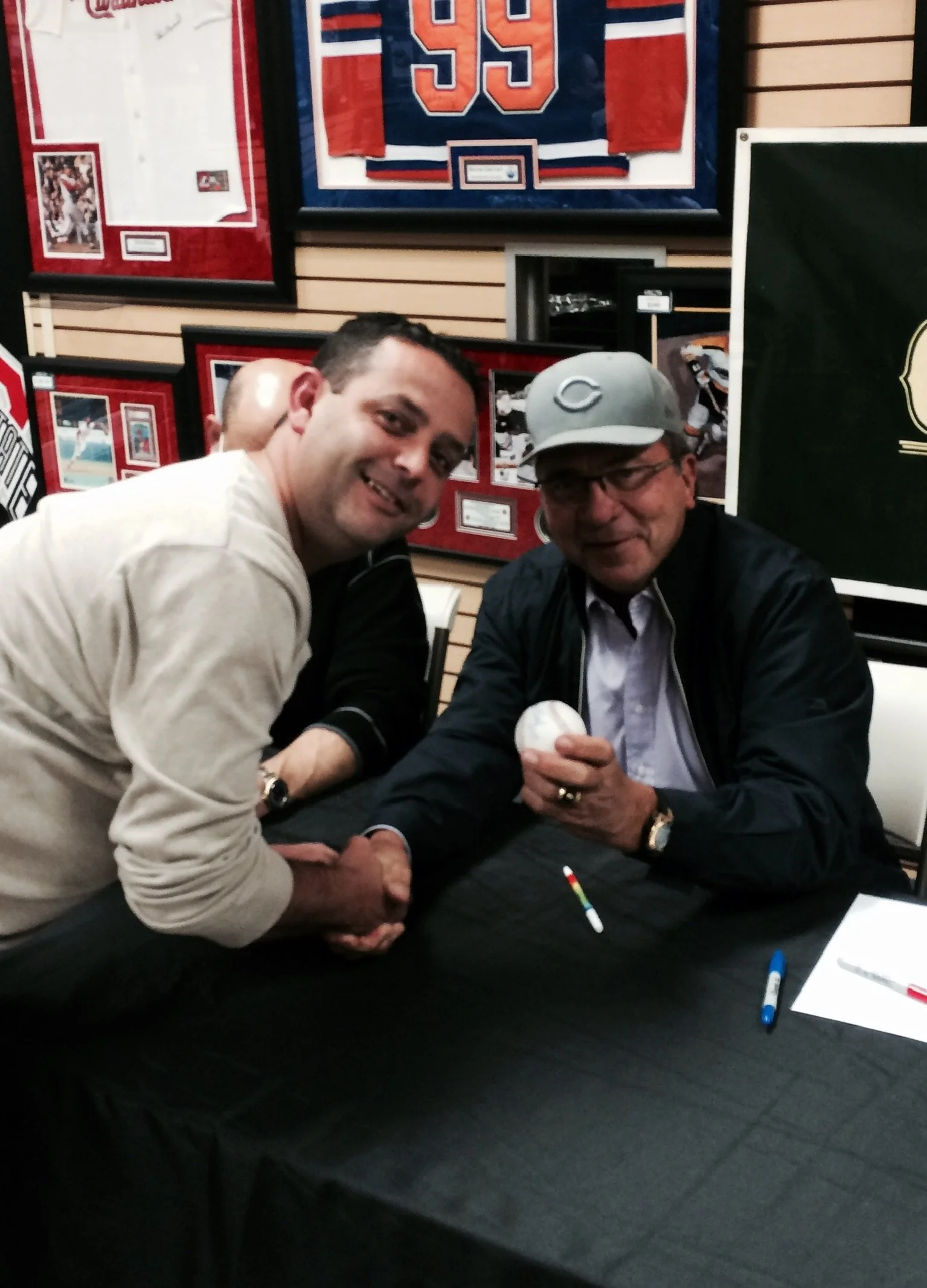 A man in a beige sweater shakes hands with a man in a baseball cap and glasses who is holding a baseball, seated at a black table with markers and a paper, in a room decorated with framed sports memorabilia.