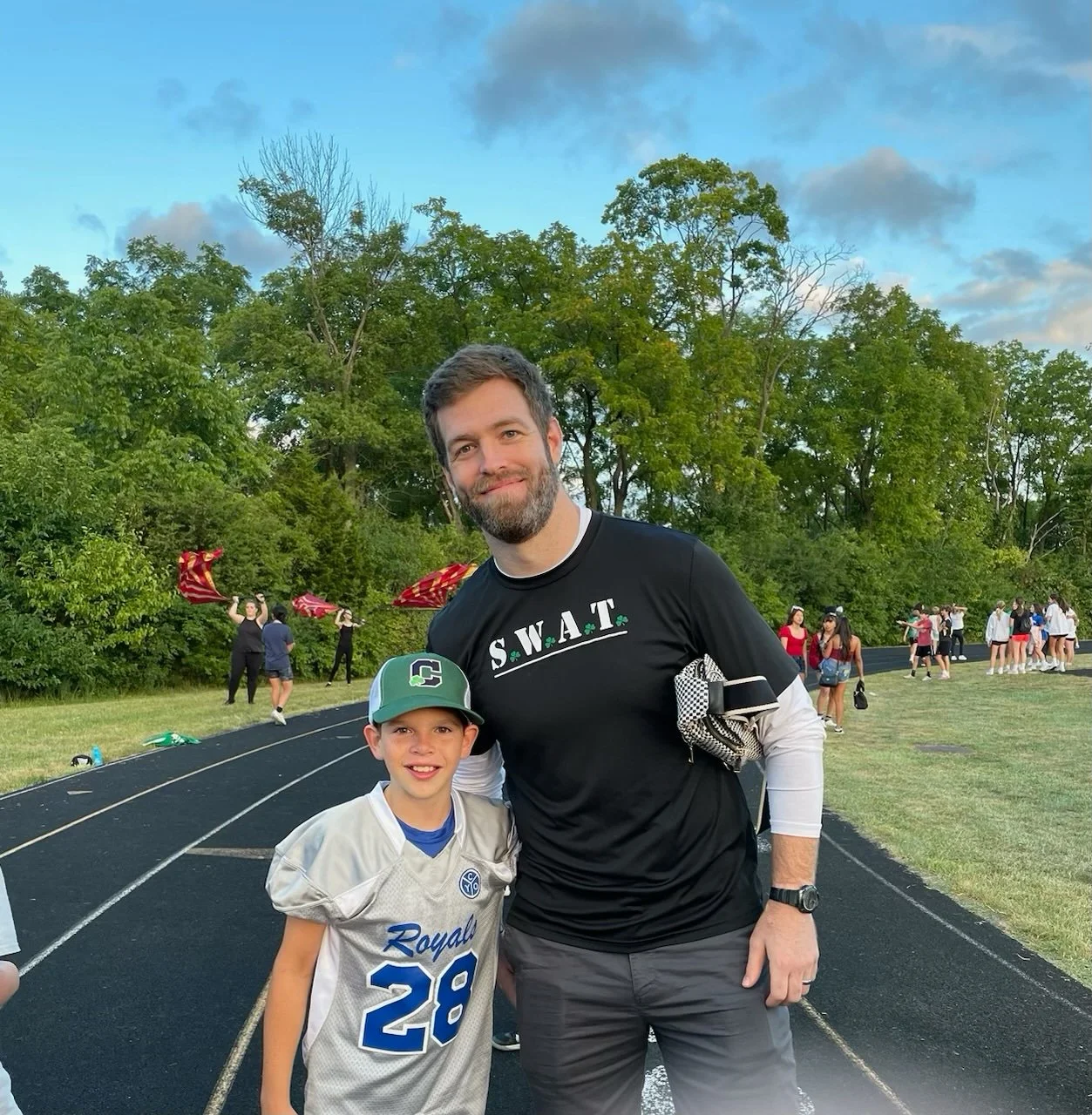 A man and a young boy standing on a running track at an outdoor sports event, with other people and trees in the background, during the daytime.