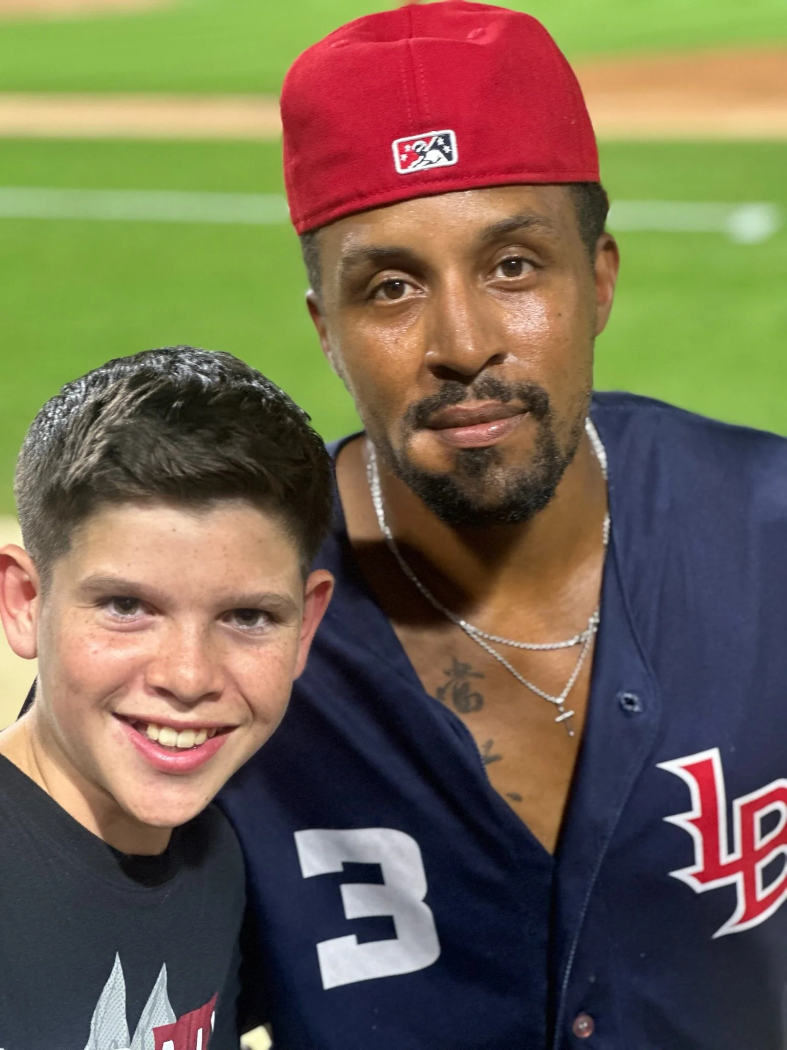 Two males taking a close-up selfie on a baseball field: one young smiling, wearing a black T-shirt, and the other adult with a beard, wearing a red cap, navy jersey with the number 3, and necklaces.