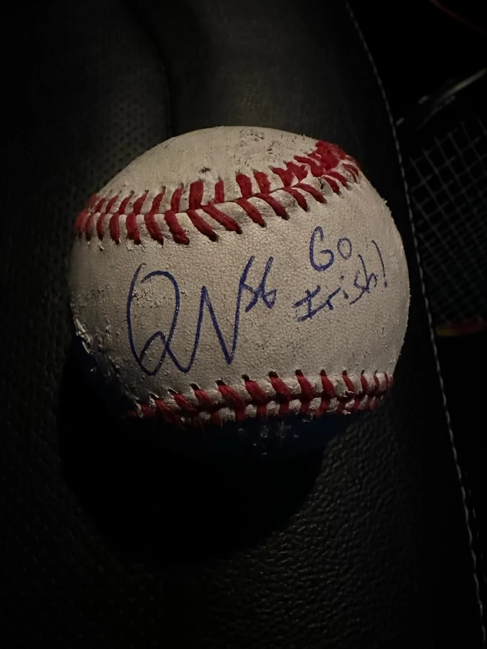 A baseball with red stitching and a handwritten message in blue ink reading 'DAN 96 GO INJ' resting on black textured material, possibly a seat.
