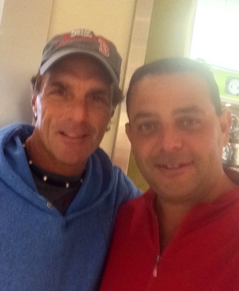 Two men taking a selfie indoors, one wearing a blue hoodie and cap, the other in a red hoodie, both smiling.