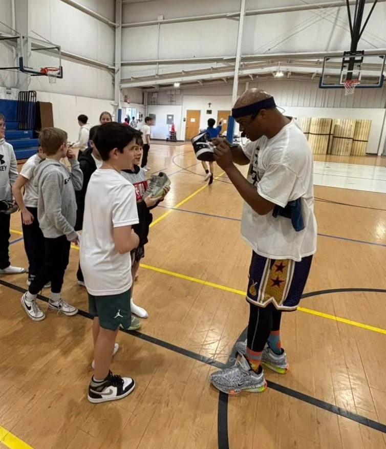 A young man wearing athletic clothing and glasses is posing to take a selfie with a group of children in a gymnasium, with other kids in line in the background.