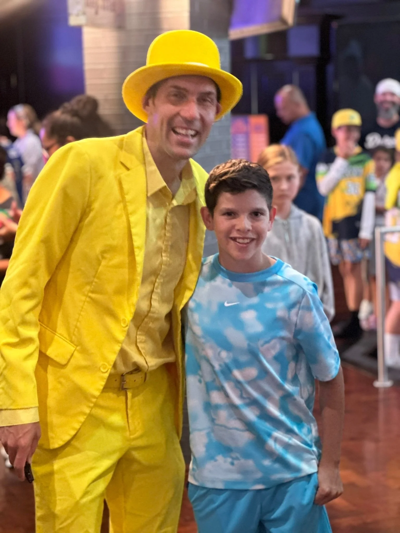 A man dressed in a bright yellow suit and hat, smiling with a young boy wearing a blue and white cloud-patterned shirt, at an indoor event with other children and adults in the background.