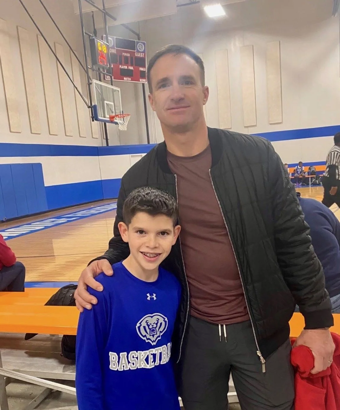 A man and a boy posing together in a basketball gym. The boy is wearing a blue basketball jersey and the man is wearing a black jacket. They are smiling and standing close together.