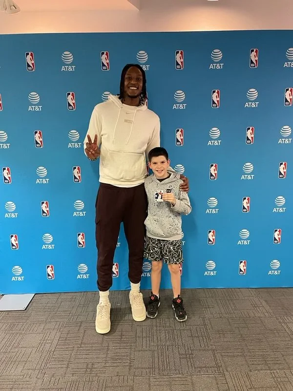 A tall man with long hair and a hoodie standing next to a young boy with short hair, both smiling, at an NBA and AT&T backdrop, with the man making a peace sign and the boy holding a small item.