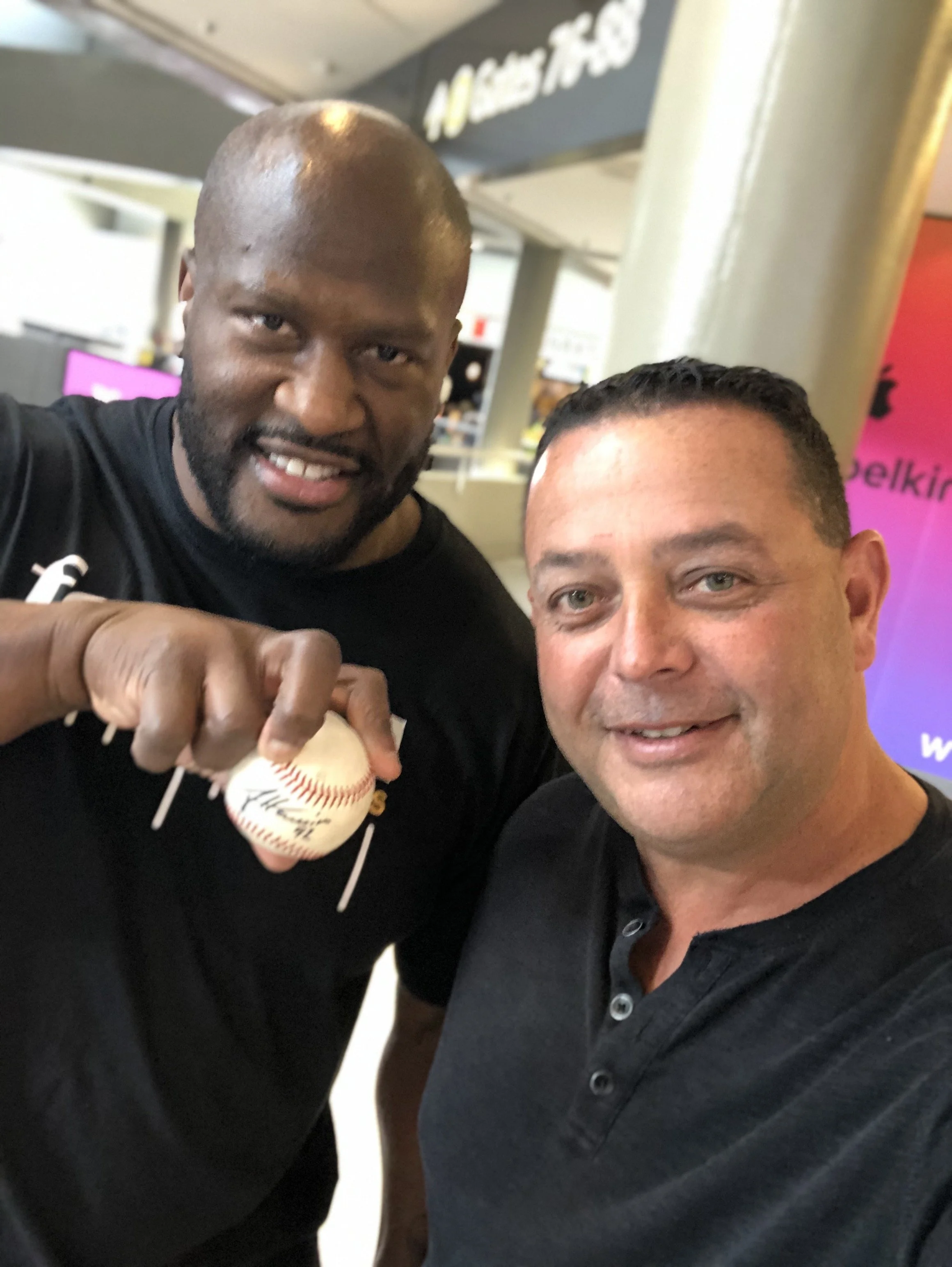 Two men taking a selfie, one holding a baseball, in an indoor setting.
