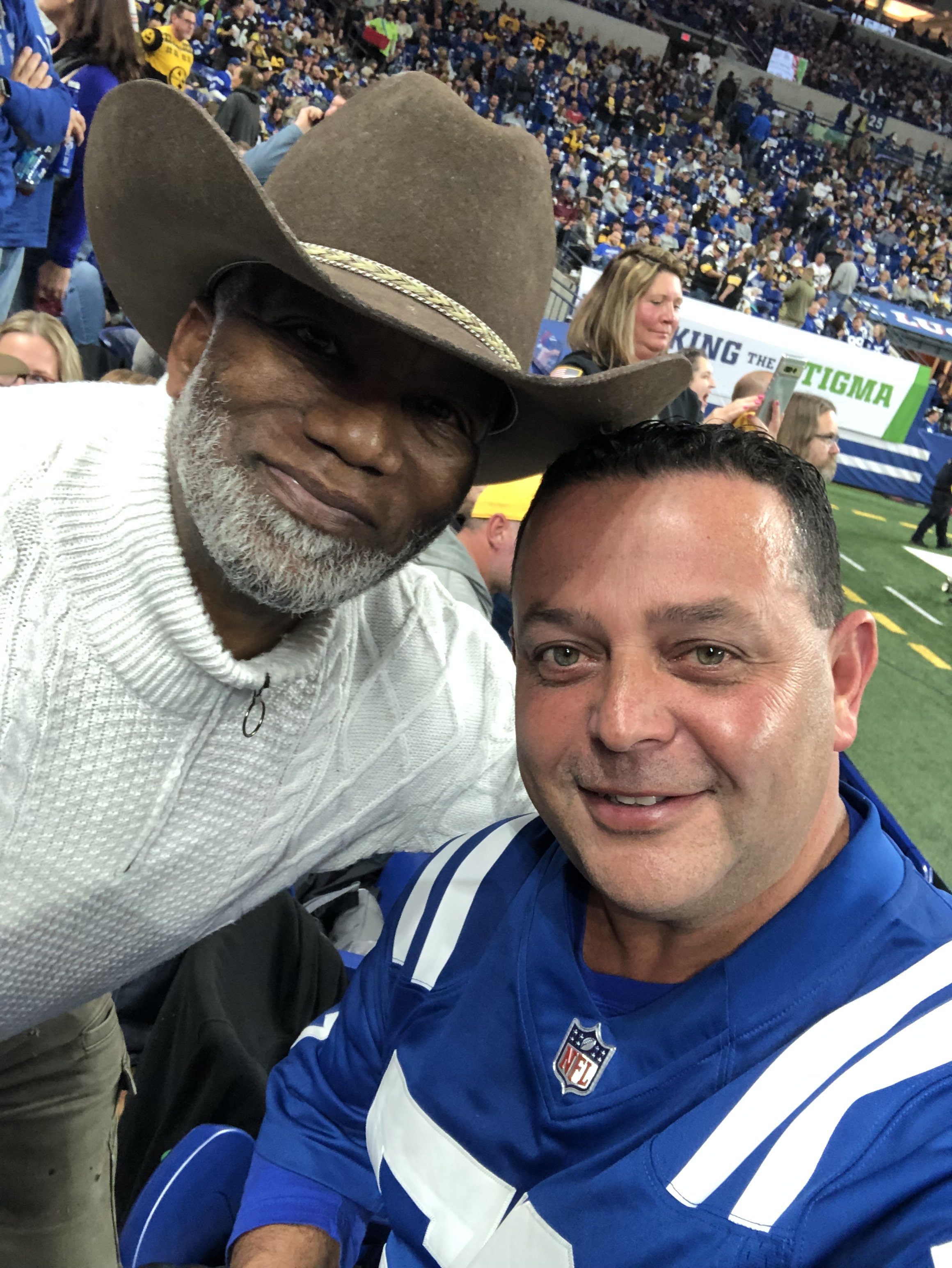 Two men taking a selfie at a stadium during a football game, with a crowd in the background. One man wears a cowboy hat and a white sweater; the other wears a blue football jersey.