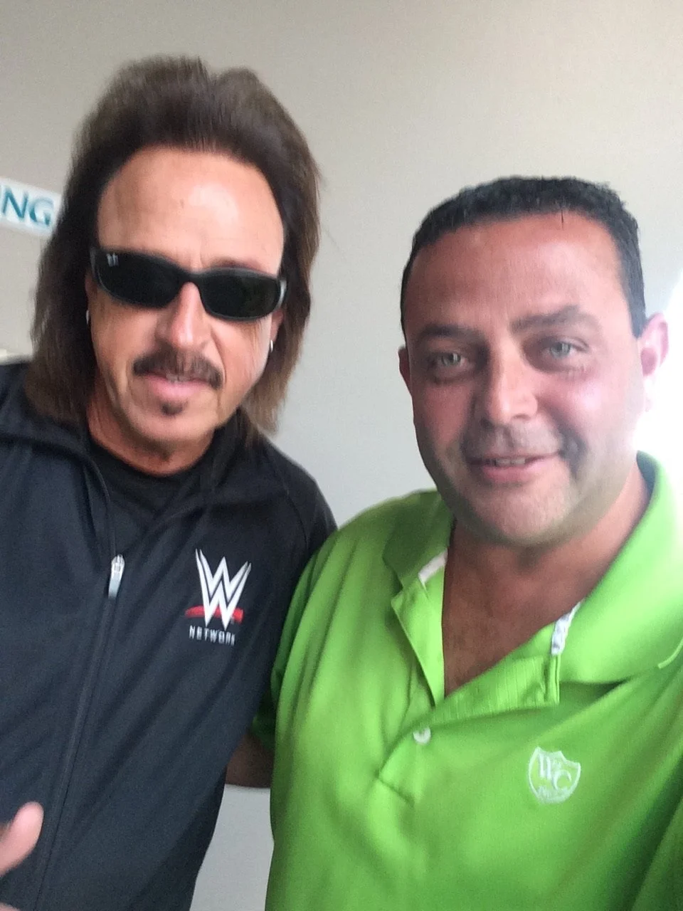 Two men posing for a photo, one is wearing sunglasses and a black WWE Network jacket, and the other is in a bright green polo shirt. Both are smiling at the camera.