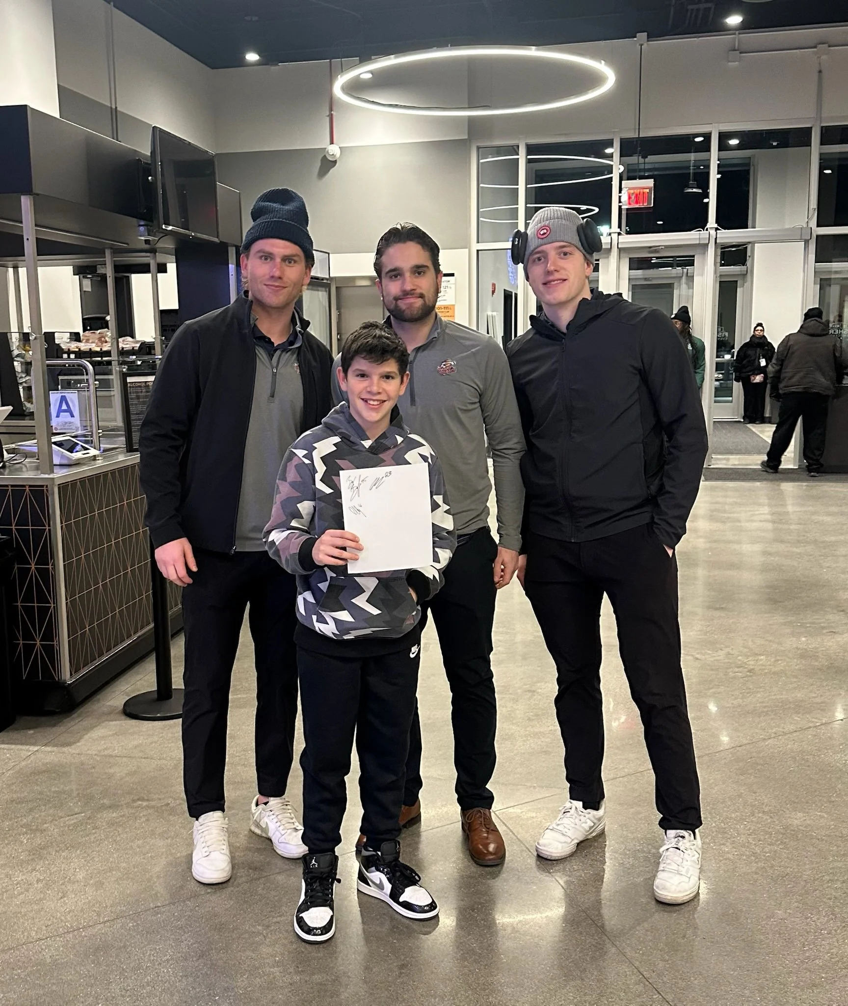 A group of four men and one boy standing inside a modern building, possibly an airport or theater, with some people in the background. The boy is holding a piece of paper or a booklet, and smiling at the camera.