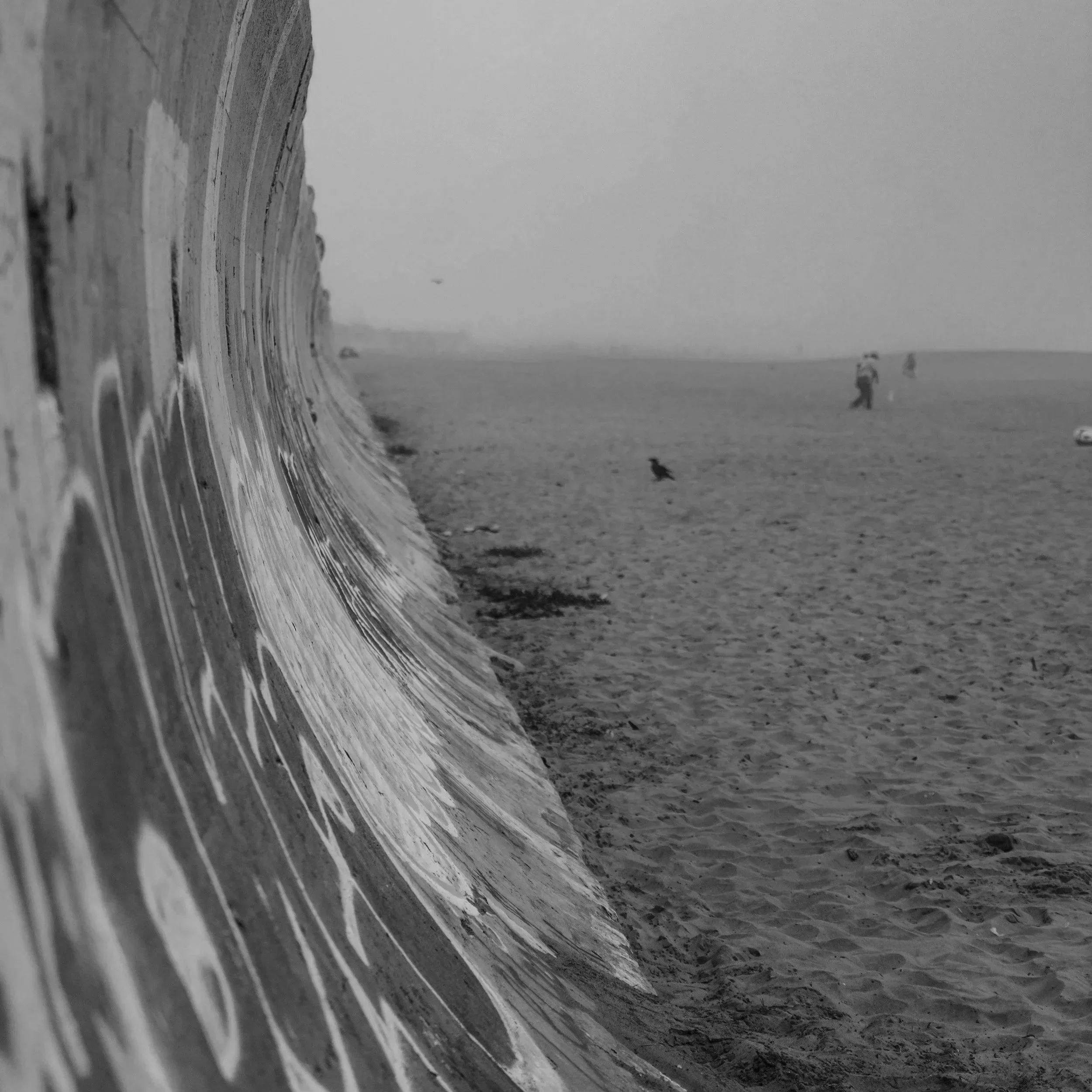A black-and-white photograph of a sandy beach with a high wooden seawall on the left side, and a few people walking or standing in the distance. The sky is overcast and the scene appears to be calm and quiet.