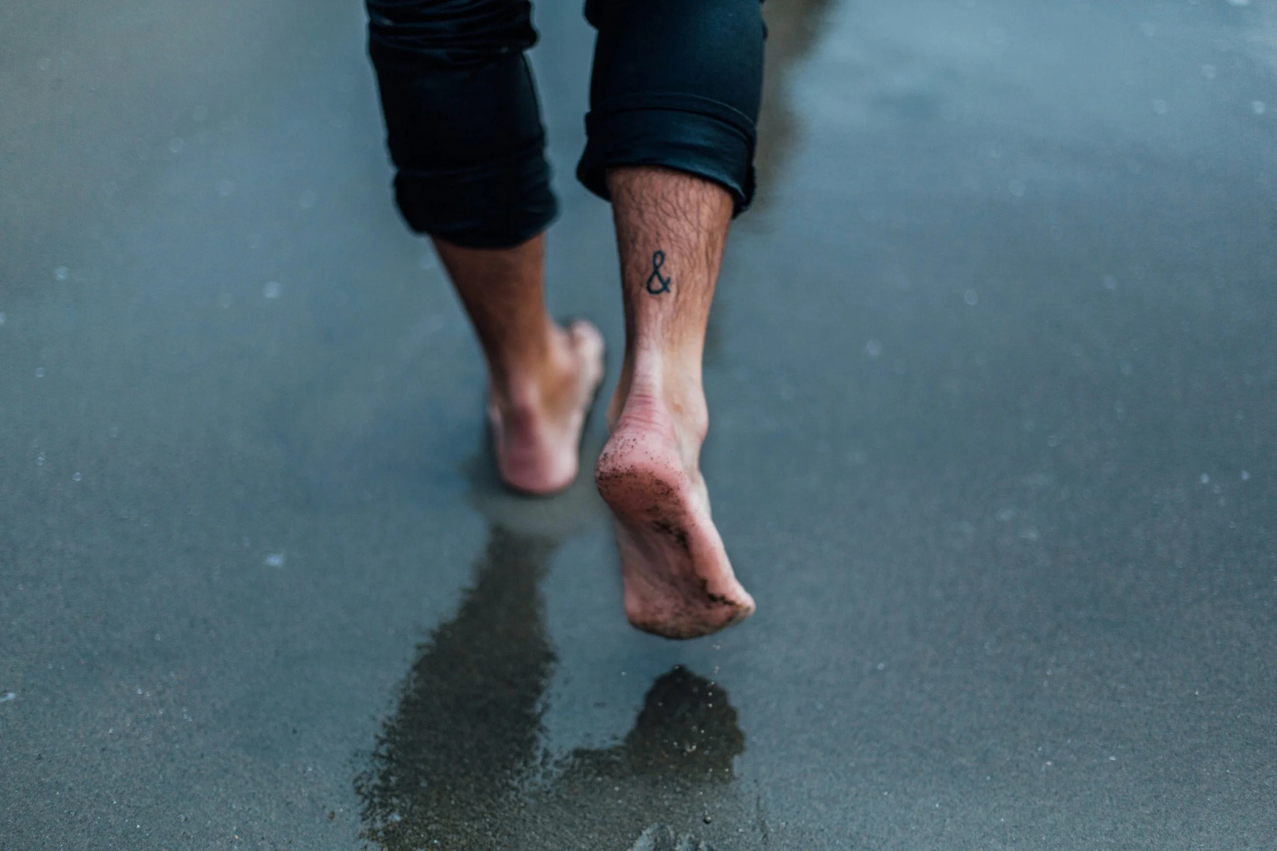 A person with rolled-up black pants reveals a small tattoo of an ampersand on their right calf. They are walking barefoot on a wet surface, and the reflection of their feet can be seen on the ground.