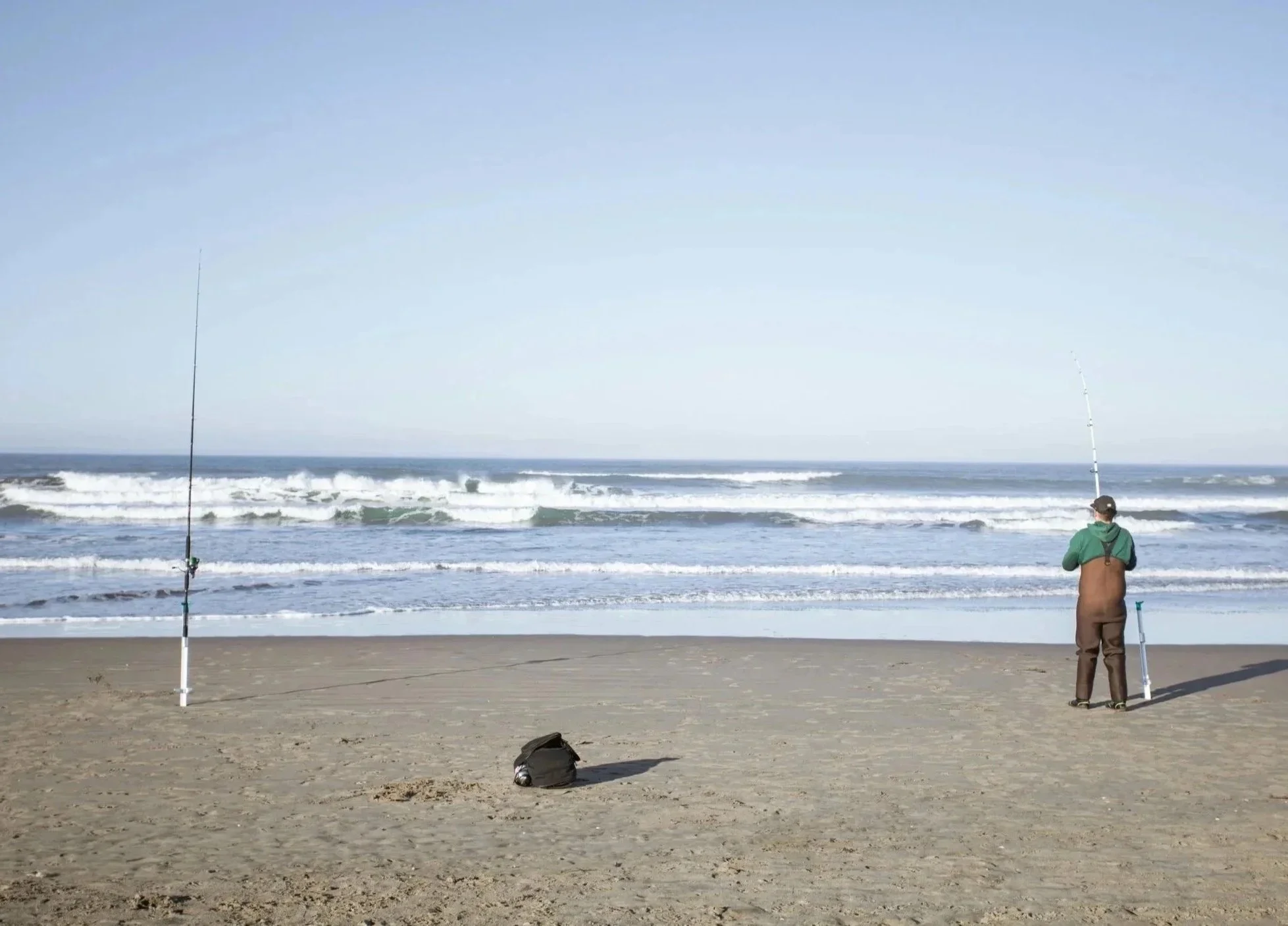 Beach scene with two fishing poles set up on the sand, a person standing near the water with two fishing rods, wearing a hoodie and cap, and a black backpack on the sand.