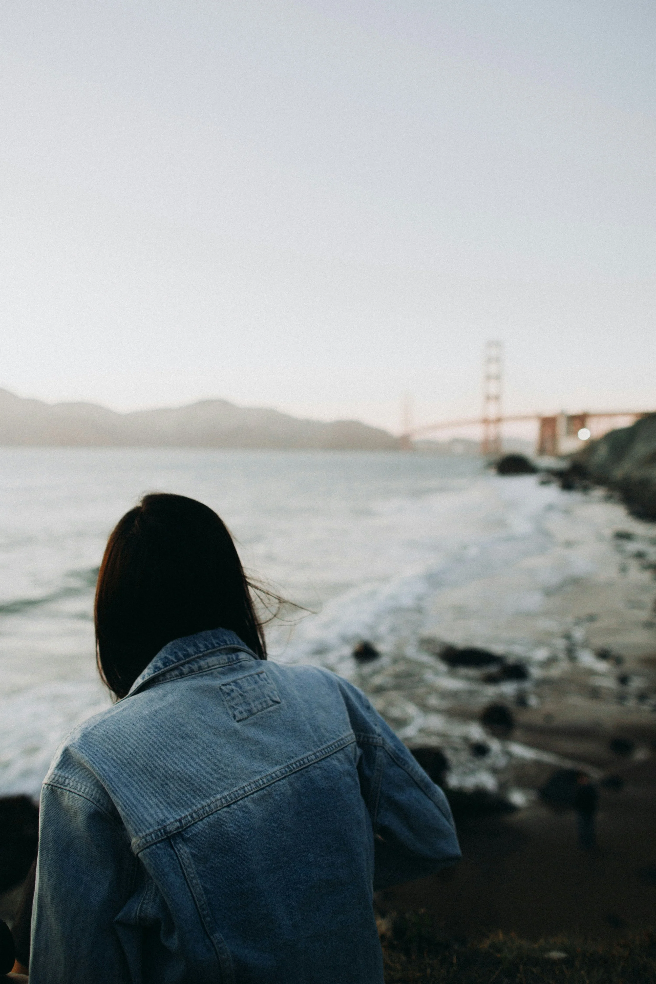 Person sitting on a rocky beach facing the water with the Golden Gate Bridge visible in the distance during dusk.