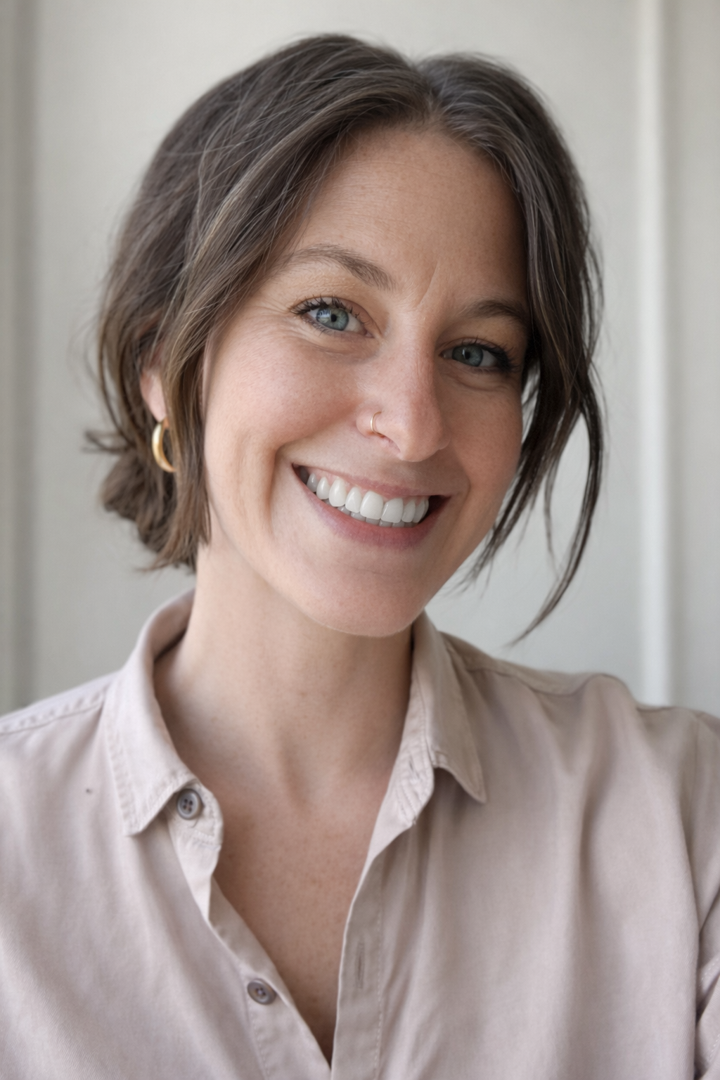 Close-up of a smiling woman with short brown hair, wearing a beige button-up shirt and gold hoop earrings, standing against a neutral background.