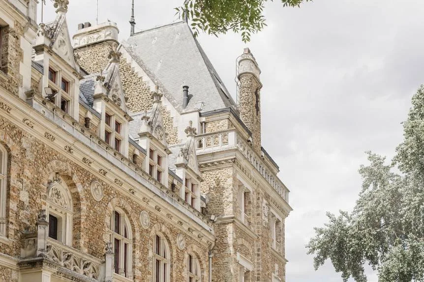 Historic stone building with ornate architectural details, multiple windows, and a tower, surrounded by trees, under a cloudy sky.