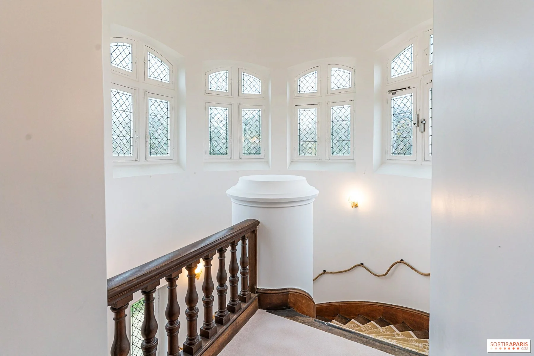 View of an interior staircase landing with tall, arched windows with diamond-patterned glass. The area has white walls, a wooden handrail, and a small decorative wall light.