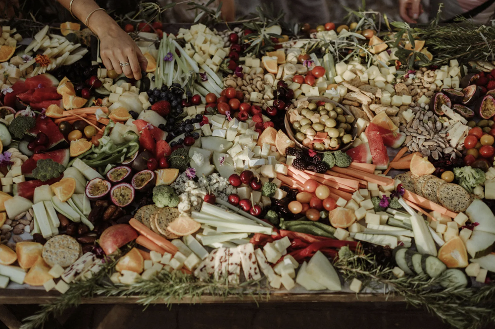 A large platter of assorted cheeses, fruits, vegetables, and crackers garnished with greenery, with a person's hand reaching for food.