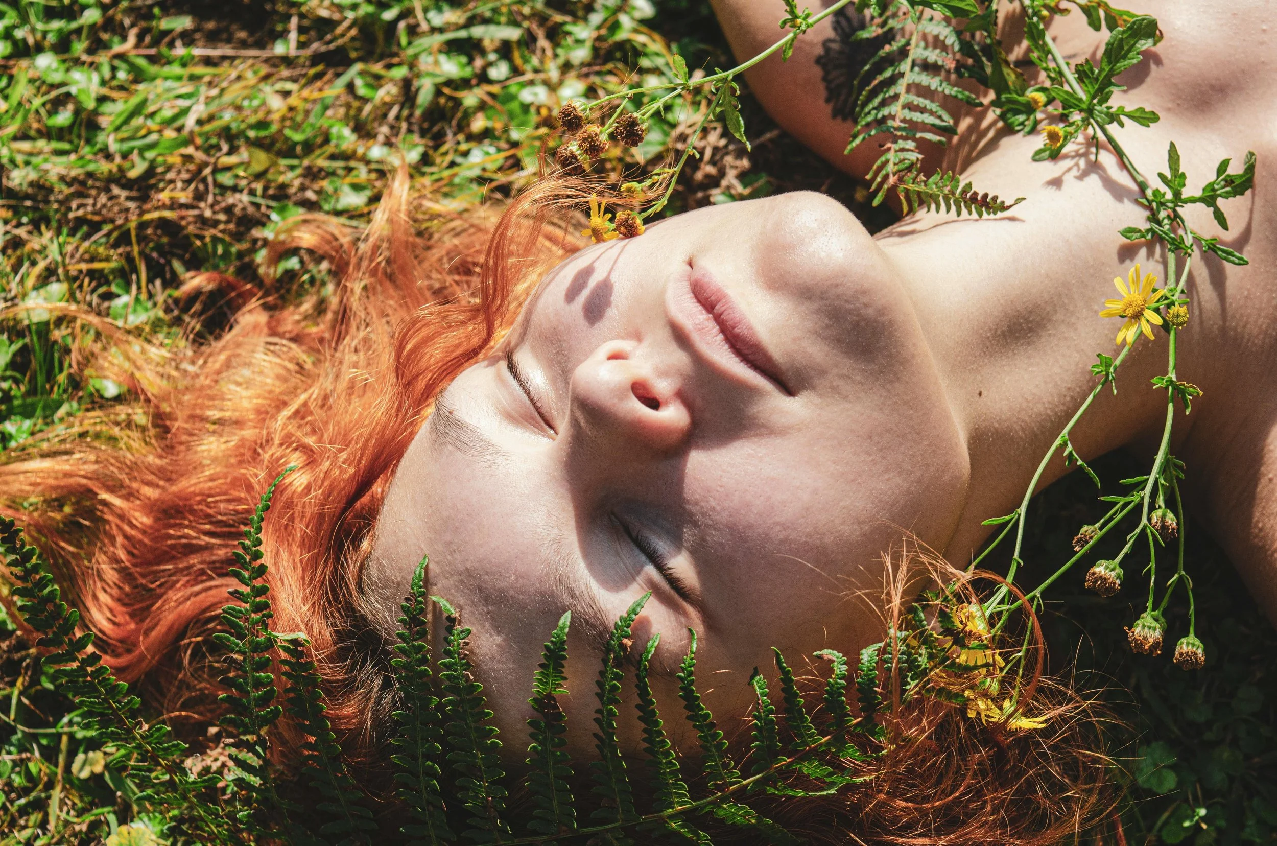 A woman with curly red hair lying on the ground outdoors, surrounded by greenery and yellow wildflowers, with her eyes closed.