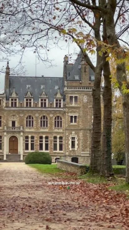A historic stone mansion with large windows and a steeply pitched roof, surrounded by trees with autumn leaves, some fallen on the ground.