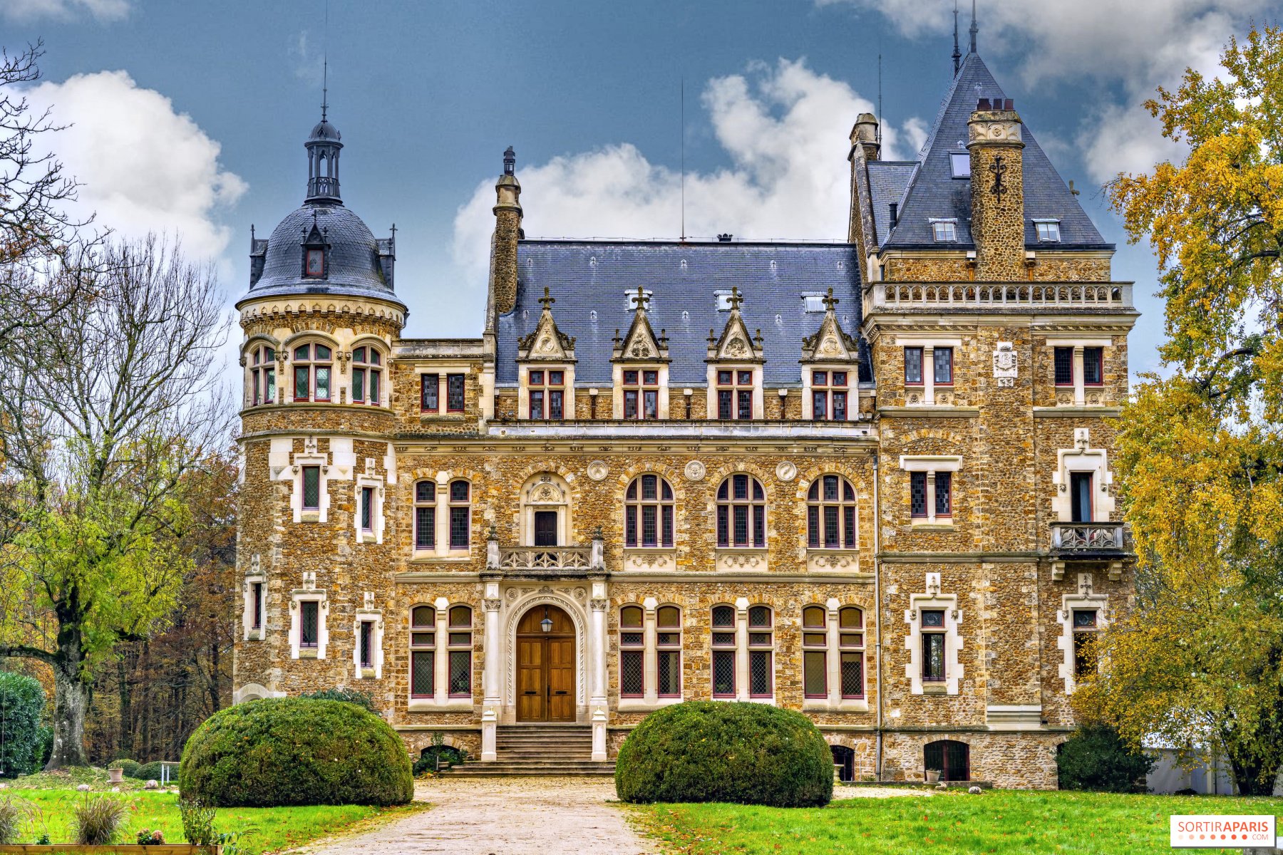 Front view of a historic stone castle with multiple towers, arched windows, and a wooden door, surrounded by trees with some leaves changing color.