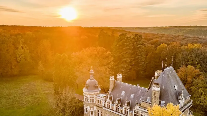 A historic castle with turrets and a steep roof is surrounded by lush autumn trees, with the setting sun casting a golden glow in the distance.